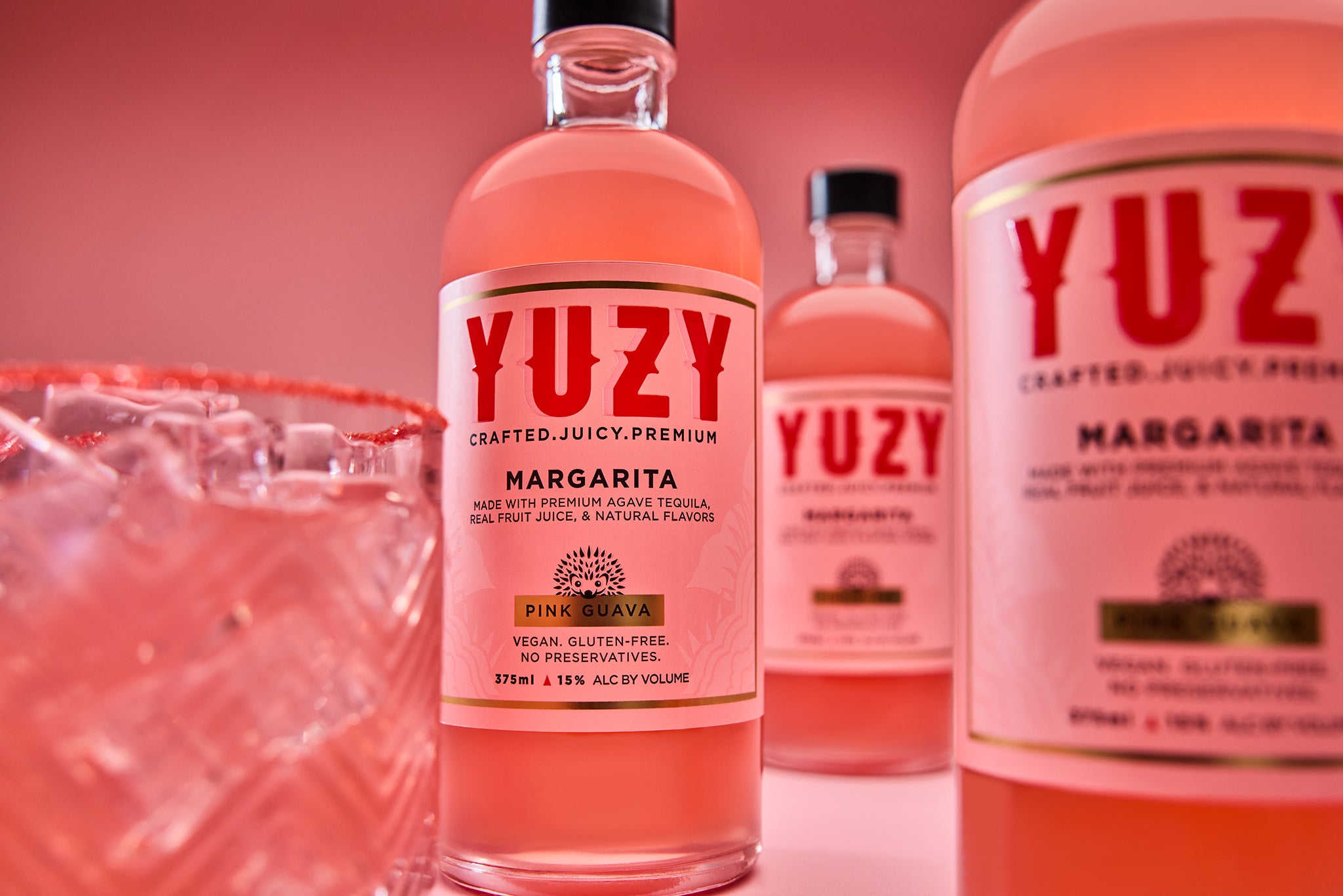 Bottles of YUZY "Pink Guava" Margaritas and a Margarita in a cocktail glass, on the rocks or on ice, on a pink background.